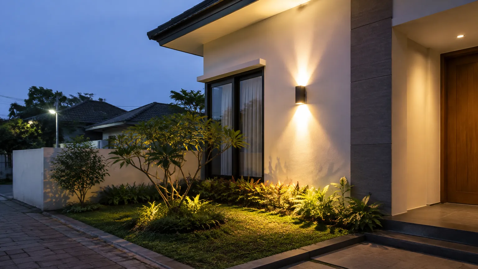 Lampu dinding menerangi fasad rumah dan tanaman di halaman saat senja