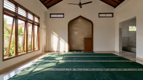 Ruang musala dengan sajadah hijau, jendela, dan kipas plafon