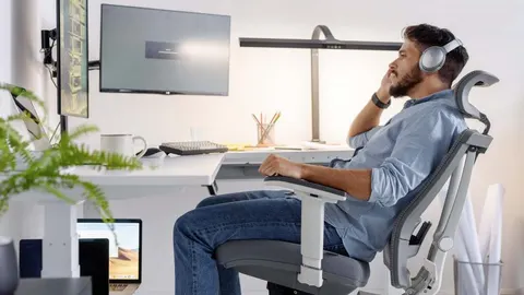 Pria duduk di kursi kantor bersandaran kepala di depan meja dan layar komputer.
